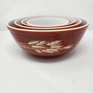 May include: A set of four brown mixing bowls, nested together. Each bowl has a white interior and a cream-coloured wheat stalk design. The bowls are arranged in a stack, showing their different sizes. The bowls are in a warm brown colour.