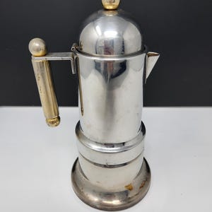 Puede incluir: Una cafetera italiana de acero inoxidable con un asa y una parte superior doradas. La cafetera tiene una base redonda y una tapa abovedada con un pomo dorado. El asa está fijada al lateral de la cafetera.