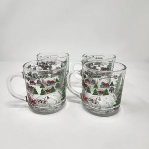 May include: A set of four clear glass mugs featuring a wrap-around winter scene. The design includes red houses, green pine trees, and a white horse-drawn sleigh with a red carriage. Each mug has a curved handle and a smooth, transparent finish.