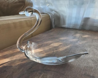 May include: A clear glass swan-shaped decorative bowl. The swan has a long, curved neck and a wide, shallow bowl for the body. The glass is transparent, allowing light to pass through. The bowl is placed on a wooden surface.