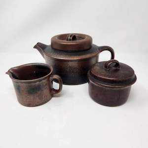 May include: A dark brown ceramic tea set consisting of a teapot with a lid, a creamer pitcher, and a lidded sugar bowl. Each piece features a mottled, textured finish with darker blackish-brown accents and a smooth, rounded handle design.