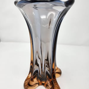 May include: A tall, elegant glass vase with a flared top and a three-footed base. The vase features a gradient of colours, transitioning from a dark brown base to a light grey centre and a dark blue rim. The vase is approximately 25 cm tall.