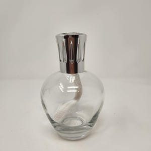 May include: A clear glass fragrance lamp with a rounded body and a thick white cotton wick inside. The top features a polished silver-coloured metal crown with vertical cutouts, designed to sit securely on the neck of the glass vessel.