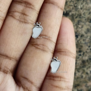Puede incluir: Dos pequeños pendientes plateados con forma de fresa. Cada pendiente presenta un centro blanco en forma de pera, un contorno plateado y un pequeño detalle de corona en la parte superior. La palabra "Springreen" está impresa en los pendientes.