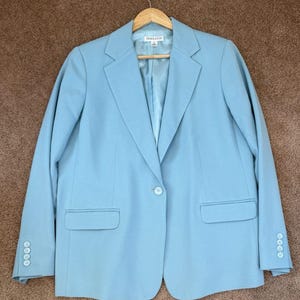 Könnte beinhalten: Hellblauer Blazer mit Ein-Knopf-Verschluss, zwei Vordertaschen und drei Knöpfen an jeder Manschette. Der Blazer hängt an einem Holzbügel. Der Markenname "Pendleton" ist auf dem Etikett zu sehen.