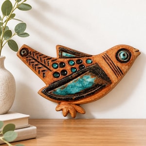 Può includere: Un ornamento da parete in ceramica a forma di uccello color terracotta con accenti smaltati turchesi, dettagli marrone scuro e un occhio circolare prominente. L'uccello presenta motivi a piuma incisi e una superficie ruvida.