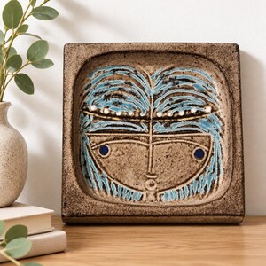 May include: A square ceramic wall plaque with a speckled brown finish. The center features a stylized face design with textured blue hair, a thin gold horizontal line, and two dark blue circular eyes set against the brown clay surface.