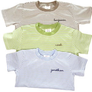 May include: Three short-sleeved t-shirts with horizontal stripes in brown, green, and light blue. Each shirt features a solid-colored crew neckline and a name embroidered in cursive script on the chest: Benjamin, Noah, and Jonathan respectively.
