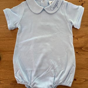 May include: A light blue short-sleeve romper featuring a rounded Peter Pan collar with the name Cooper embroidered in dark thread on the right side. The garment includes a button closure at the neckline and snap closures at the bottom hem.