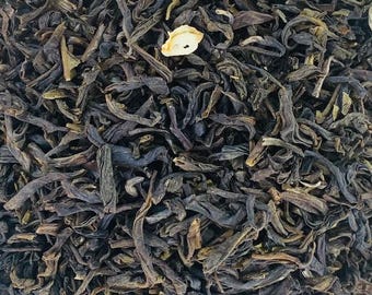 May include: Close-up of loose leaf tea with a mix of dark green and brown leaves. A single, light-colored, dried flower petal is visible among the tea leaves. The tea leaves have a slightly twisted appearance, suggesting a high-quality blend.