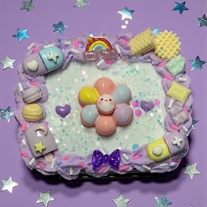 May include: A decorative phone case with a whimsical design. The case features a flower with a smiling face, a rainbow, and various miniature food items. The border is decorated with purple frosting, sprinkles, and small candies. The background is a light purple color with star-shaped decorations.
