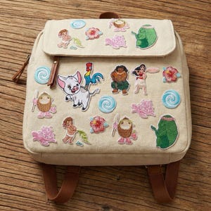 Può includere: Zaino beige con spallacci marroni e chiusura con patta. Lo zaino è decorato con toppe ricamate di personaggi e simboli di un popolare film d'animazione, tra cui un maiale, un gallo e vari fiori tropicali.