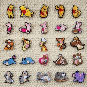 Puede incluir: Una colección de coloridos dijes de personajes de Winnie the Pooh. Los dijes presentan varios personajes, incluyendo a Winnie the Pooh, Piglet, Tigger y Eeyore, en diferentes poses. Los dijes están colocados sobre un fondo neutro y texturizado.