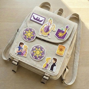 Op de afbeelding: Een lichtgrijze rugzak versierd met kleurrijke patches met personages en symbolen uit de animatiefilm Rapunzel. Patches zijn onder meer een boot, een zon en personages met lang blond haar.