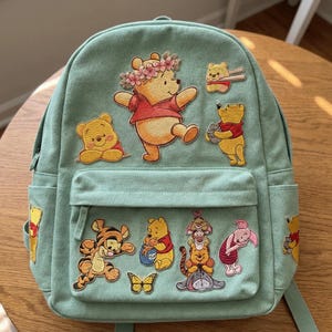 May include: A light green backpack featuring multiple Winnie the Pooh character patches. The patches include Winnie the Pooh, Tigger, Piglet, and Eeyore. The backpack has a front pocket and a top handle.
