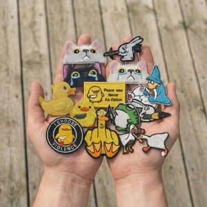 May include: A collection of embroidered patches featuring various designs. Included are cat faces, ducks, a frog, a wizard, and a rabbit. Some patches have text that reads "Peace was Never An Option" and "I Choose Violence".