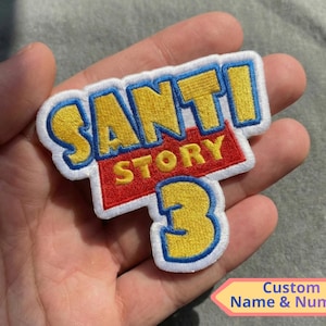 May include: Embroidered patch with the text "SANTI STORY 3" in yellow and blue, on a white background. The word "STORY" is on a red background. The patch is held in a hand. The bottom right corner has a small sign that says "Custom Name & Number".