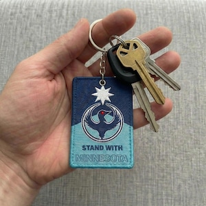Può includere: Un portachiavi rettangolare con un design blu e turchese, con un'emblema di uccello e la scritta "STAND WITH MINNESOTA". Il portachiavi è attaccato a un anello di metallo che tiene diverse chiavi.