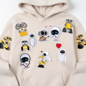 Puede incluir: Sudadera con capucha beige adornada con varios parches bordados con personajes de la película animada Wall-E. Los parches incluyen robots en amarillo, blanco y gris, con detalles de corazones.