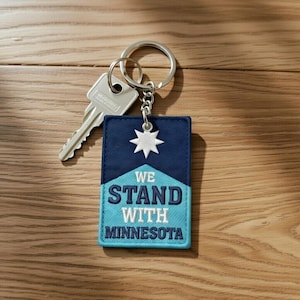 Puede incluir: Un llavero rectangular con una llave y un anillo plateados. El llavero tiene una parte superior azul marino con una estrella blanca y una parte inferior azul claro con el texto "WE STAND WITH MINNESOTA".