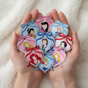 May include: A collection of heart-shaped pins featuring various cartoon princess characters. Each pin has a pearl border, a bow, and a portrait of a princess. The pins are in shades of pink, blue, and red.