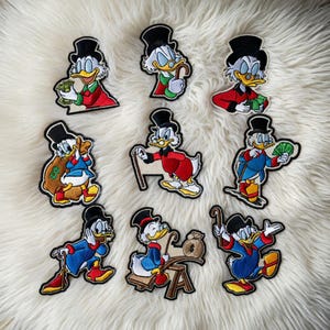 May include: A collection of nine embroidered patches featuring Scrooge McDuck in various poses. The patches showcase Scrooge in a black top hat, red coat, and blue suit, holding money, a cane, and a money bag. The patches are on a white, fluffy surface.