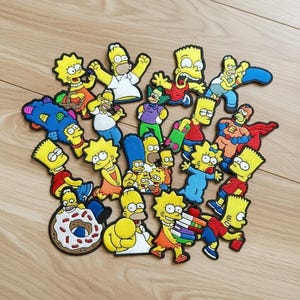 May include: A selection of embroidered patches featuring characters from The Simpsons. The patches show Homer, Marge, Bart, Lisa, and other characters in various poses, with bright yellow skin and colourful clothing.