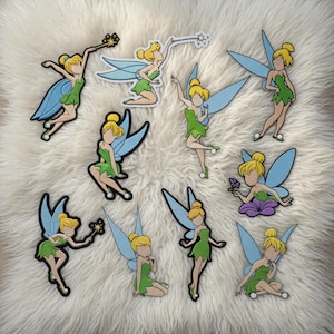 Puede incluir: Una colección de nueve parches de hadas de dibujos animados. Cada parche presenta un hada rubia con alas azules y un vestido verde. Algunas hadas sostienen varitas o flores. Los parches están sobre una superficie blanca y esponjosa.