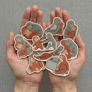 Könnte beinhalten: Eine Sammlung von aufgestickten Aufnähern mit verschiedenen Affenmotiven in Grau-, Orange- und Cremetönen. Die Aufnäher zeigen Affen in verschiedenen Posen, darunter Umarmen, Sitzen und Interagieren. Die Aufnäher werden in offenen Händen gehalten.