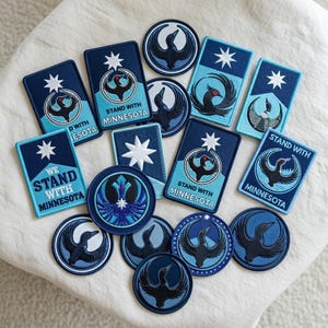 May include: A collection of navy blue and light blue embroidered patches. The patches feature a stylized bird design, a star, and the text "STAND WITH MINNESOTA" or "WE STAND WITH MINNESOTA".