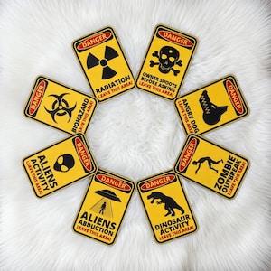 May include: A collection of yellow danger signs with black graphics and red borders. The signs warn of radiation, biohazards, alien activity, alien abduction, angry dogs, dinosaur activity, and zombie outbreaks. Each sign includes the text "Danger" and "Leave this area!"