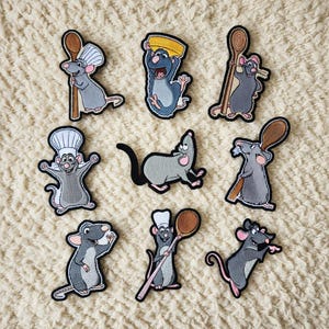 Può includere: Una collezione di nove toppe ricamate raffiguranti topi dei cartoni animati in varie pose. I topi sono principalmente grigi con orecchie e zampe rosa, alcuni indossano cappelli da chef o tengono utensili da cucina. Le toppe hanno contorni neri e sono su sfondo crema.