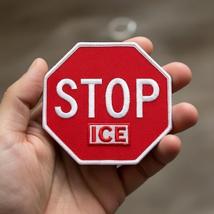 Op de afbeelding: Een rode, achthoekige patch met witte geborduurde tekst "STOP" boven "ICE". De patch heeft een witte rand en wordt in een hand gehouden. De patch lijkt op een stopbord.