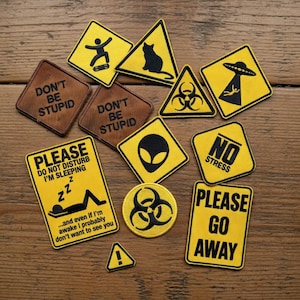 Może przedstawiać: Zbiór żółto-czarnych naszywek z różnymi wzorami i tekstami, w tym ze skaterem, kotem, obcym i napisami "DON'T BE STUPID" i "PLEASE GO AWAY". Naszywki są ułożone na drewnianej powierzchni.