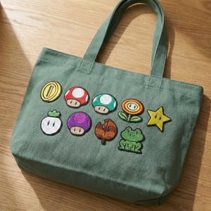 May include: A green canvas tote bag featuring embroidered patches of various Super Mario Bros. characters and icons. The patches include a gold coin, mushrooms, a flower, a star, and a frog. The bag has two handles and is sitting on a wooden surface.