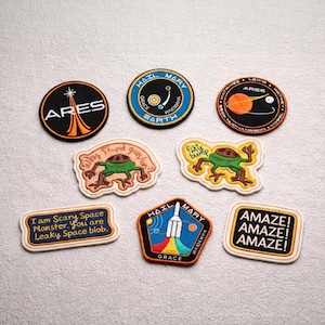 May include: A collection of embroidered patches with various designs and text. The patches feature space-themed graphics, including rockets, planets, and the word "ARES." Some patches have phrases like "AMAZE!" and "I am Scary Space Monster."