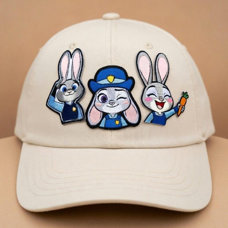 May include: A beige baseball cap featuring three embroidered patches of a cartoon rabbit character. The patches depict the rabbit in different poses, wearing a police uniform and holding a carrot. The cap has a curved brim and a button on top.