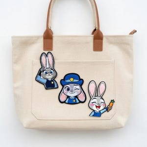 May include: Beige canvas tote bag with brown leather handles and three embroidered patches of a bunny character. The patches depict the bunny in different poses, wearing a police uniform and holding a carrot.