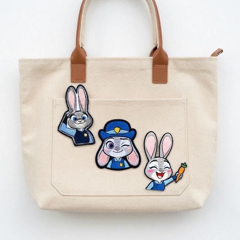 May include: Beige canvas tote bag with brown leather handles and three embroidered patches of a bunny character. The patches depict the bunny in different poses, wearing a police uniform and holding a carrot.