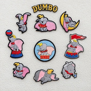 Könnte beinhalten: Eine Sammlung von gestickten Aufnähern mit der Figur Dumbo. Die Aufnäher zeigen Dumbo in verschiedenen Posen, mit rosa Ohren, einem gelben Hut und einem roten Kragen. Das Wort "DUMBO" ist auf einigen Aufnähern gestickt.