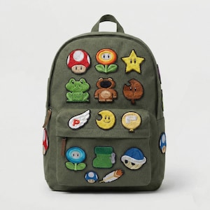 May include: Olive green backpack with embroidered patches of various cartoon characters. The patches include a red and white mushroom, a yellow star, a green frog, and a brown bear. The backpack has a front pocket and a top handle.