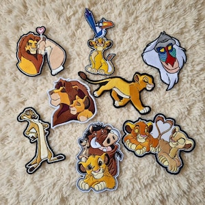 Op de afbeelding: Een verzameling geborduurde patches met personages uit de film The Lion King. De patches tonen Simba, Mufasa, Timon, Pumbaa en Rafiki, in verschillende poses en scènes. De patches zijn in tinten geel, bruin en beige.