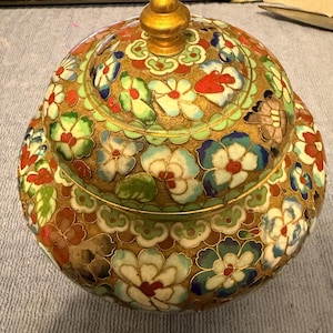 May include: A decorative, round, lidded jar with a gold-colored base and lid. The jar is adorned with colorful floral patterns in red, white, blue, and green. The lid is topped with a gold finial.