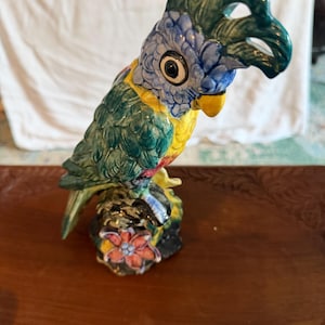 May include: A ceramic parrot figurine featuring a vibrant blue head with a dark green crest, a yellow chest, and green wings. The bird is perched on a textured base decorated with a small orange flower, showcasing a glossy finish throughout.