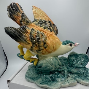 May include: A ceramic bird figurine with outstretched wings, perched on a cluster of green leaves. The bird features a mix of brown, orange, and black feathers, with a white chest and a teal head. The figurine is a decorative item.