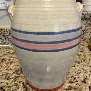 May include: A large, cylindrical ceramic crock featuring a speckled off-white glaze with horizontal bands of blue and pink. The vessel has a wide, unglazed terracotta-colored rim and base, with two small, integrated handles on the upper sides.