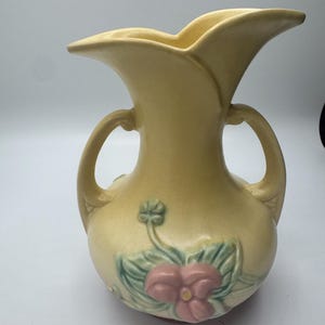 May include: A pale yellow ceramic vase with two handles and a flared, scalloped rim. The vase features a floral design with pink flowers and green leaves. The vase is approximately 8 inches tall.