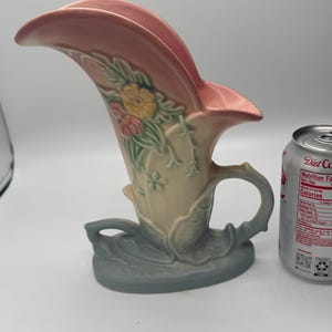 May include: A vintage art pottery vase in a stylized floral design. The vase features a pink, curved top, a cream-colored body with floral accents, and a gray-blue base. The vase has a handle and is approximately 8 inches tall.