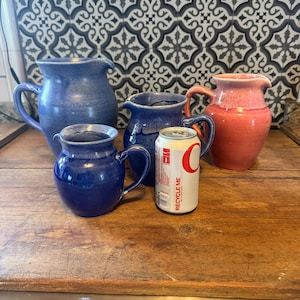 May include: A collection of ceramic pitchers in various sizes and shades of blue and coral. The pitchers have handles and spouts. A can with the text "RECYCLE ME" is placed among the pitchers.