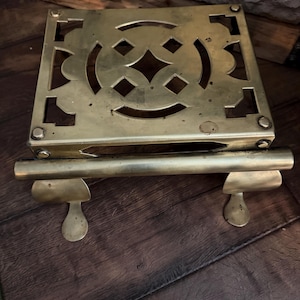 May include: A square, brass trivet with a decorative openwork design. The trivet has a handle and two small feet. The brass has a warm, aged patina. The trivet is designed to protect surfaces from hot cookware.